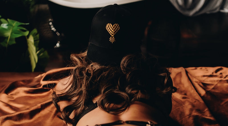 Person wearing a black cap with a gold emblem, lying on a bed with brown sheets.
